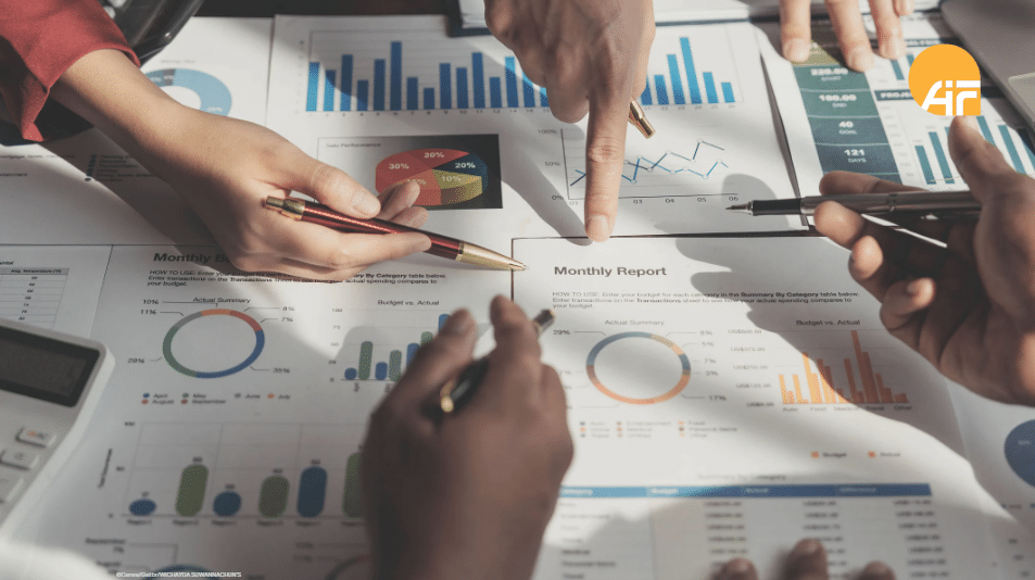 Hände mehrerer Personen zeigen auf Diagramme und Aufstellungen, die auf einem Tisch liegen.
